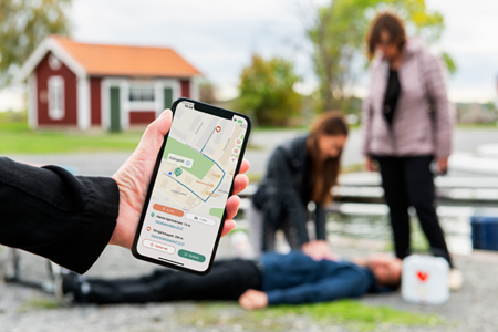 Mobil med Sms-livräddare-appen visar vägen till hjärtstartare medan personer ger hjälp vid misstänkt hjärtstopp.