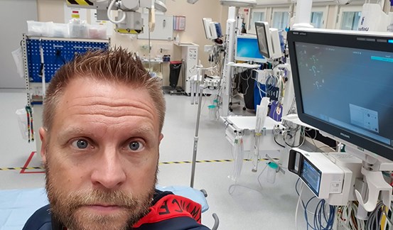 En porträttbild av Mattias Kling som står framför en  medicinteknisk apparatur med skärm och dator, apparaturen används för att ta EKG 