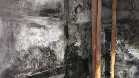 Abstrakta målningarna i svart tusch täcker väggarna i ett hörn, längst inne står tre skulpterade, bruna trädstammar.