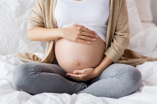 Gravid kvinna sitter med bar mage i en säng. Foto Mostphotos.