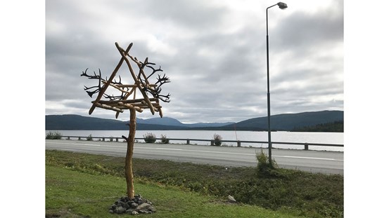 Vid vägkanten reser dig en träkonstruktion ur gräset, bortom vägen syns en sjö och fjällens linjer.
