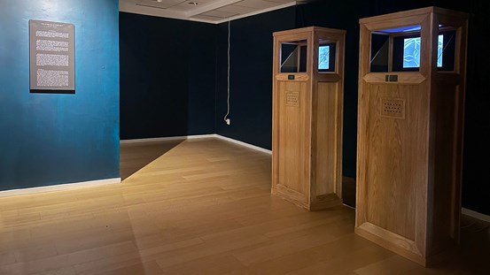 Ett utställningsrum med trägolv och djupt grönblå väggar. Till höger står två höga träskåp, montrar, som visar super 8-filmer. Till vänster en text på väggen. 