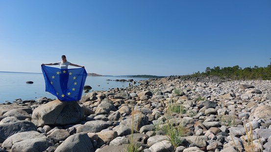 En person håller up en EU-flagga på en stenig strand vid havet.