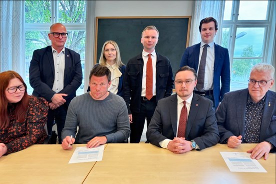 Åtta personer sitter och står bakom ett skrivbord signerar ett kontrakt