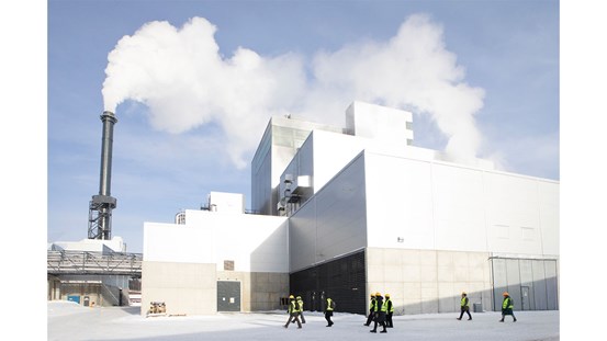 En grupp personer går framför en stor industribyggnad. Vit rök kommer ur en hög skorsten. 