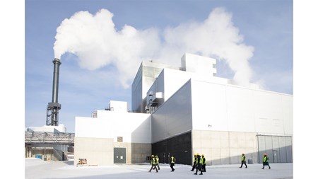 En grupp personer går framför en stor industribyggnad. Vit rök kommer ur en hög skorsten. 