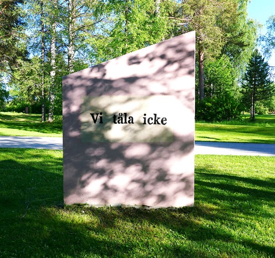 Ett ljust kantigt betongblock med en vit affisch med texten ”Vi tåla icke” i svart. Gräset runt den och blocket är lite skuggade av bladverk i träden runt om. 