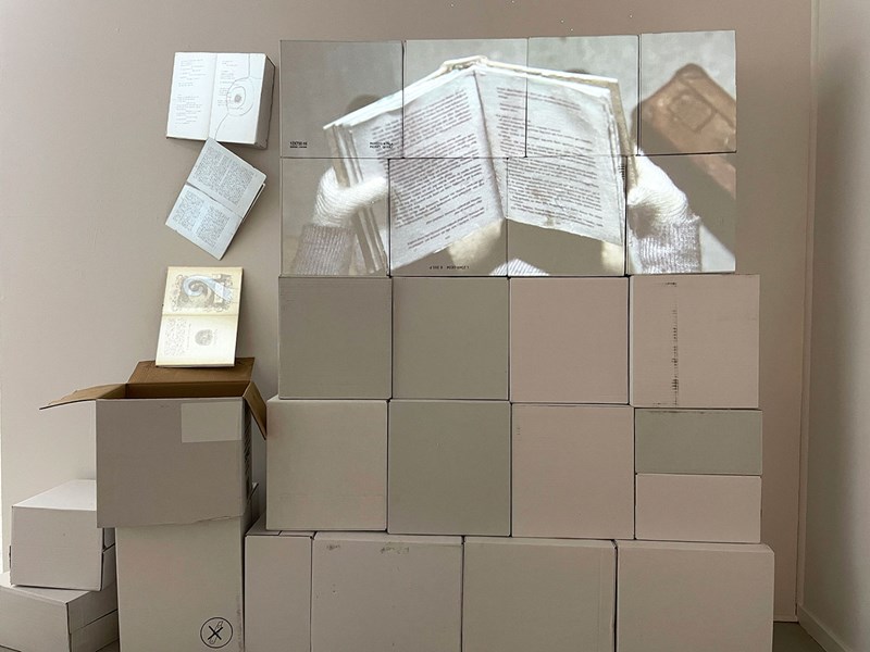 Många vita lådor står travade på varandra som en vägg och på de översta kartongerna projiceras en film. Till höger på väggen hänger böcker. På filmen visas en uppslagen bok. 