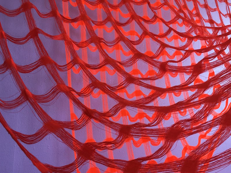 Hålgardiner där hålen mellan varptrådarna är större än den vävda ytan. Starkt röda trådar hänger ihopvävda i ett rutmönster mot en vägg i lila toner. 