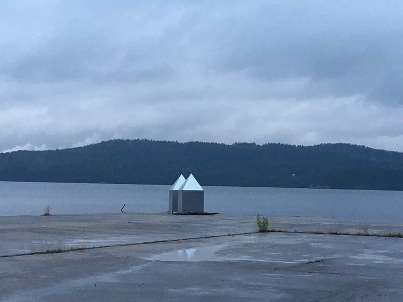 På yttersta delen av kajen fångas ljuset av en installation bestående av två järnplåts hus. De spetsiga trekantiga taken lyser som silver i den mörkgråa miljön. En pöl på betongplattan speglar de två silverfärgade taken. 