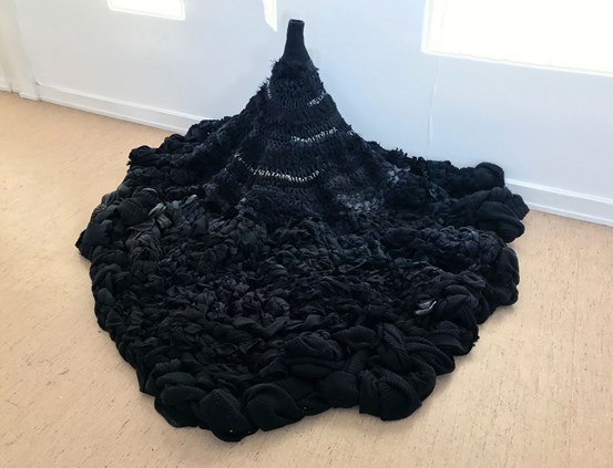 På golvet intill den vita väggen är en installation med svarta och mörkgrå textilier som arrangerats till att bilda något som liknar en vulkan, en bred bas med en smal rörliknande topp.