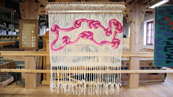 En textil bildvävnad som hänger från taket. Nästan helt transparent, skir, en gles varp där en vävd rosa, ormliknande form slingrar sig.