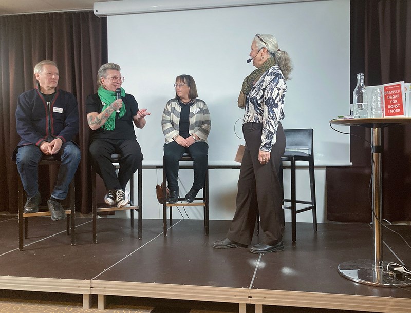 Foto. På en låg scen sitter tre personer på rad på höga stolar; Tomas, Sara och Karin. Sara håller i en mikrofon. Till höger står moderator Catarina framför en fjärde stol. Längst till höger på fotot ett högt bord med en flaska vatten och några glas. 