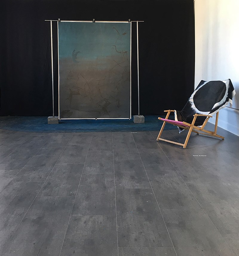 Framför ett stort svart tyg står ett screentryck uppsatt på en ställning som står på en blå yta. Ställningens ben är förankrade i vad som ser ut som betongklossar. Snett framför till höger står en solstol i trä där det ligger ett tyg med tryck på.