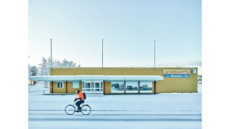Vinterdag, blå himmel. Person i orange jacka cyklar förbi. Andra sidan vägen, en gul byggnad, svartvita foton uppsatta. 