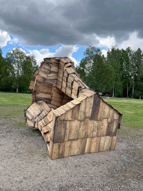 Foto. Utomhus på en grusig yta framför gräsmatta står en skulptur i trä. Gjord av takspån/pärt och består av former som avviker från förväntad geometri. 