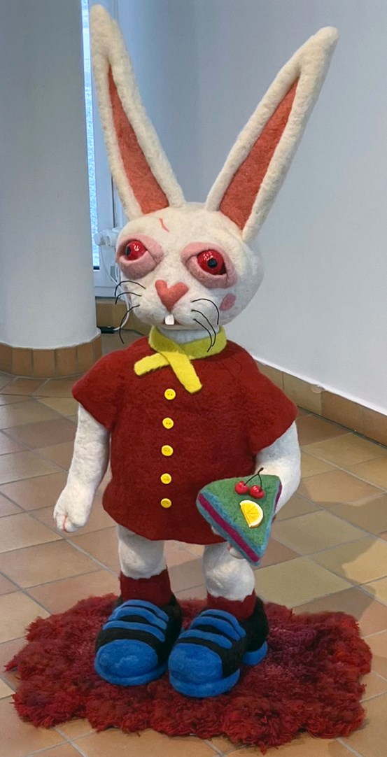 En skulptur som står på en fluffig röd matta. Det är en vit kanin med långa öron, röda ögon, röd jacka med gula detaljer och svarta och blå skor. I sin vänstra hand håller den en bit prinsesstårta. 