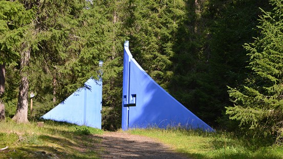 En liten gångväg leder fram till Blå porten. Två triangulärt formade, blåmålade, formgjutna betongblock står på varsin sida om stigen i solskenet. De formar en port in mot skogen. Vid sidan om stigen och bakom porten syns barrträd. 