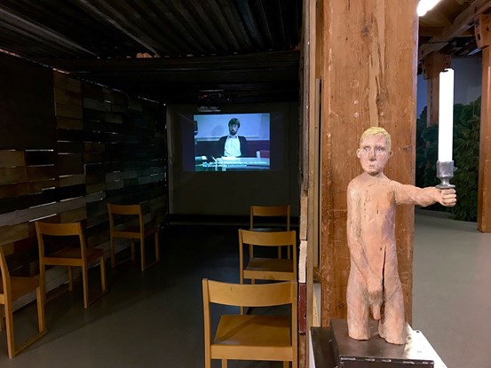 Till höger, vid ingången till ett mörklagt rum, står en träskulptur föreställande en naken, blond man hållandes i ett ljus. Längre in i rummet spelas ett videverk upp. 