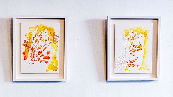 Två abstrakta bilder i främst rött och gult, i vita ramar på en vit vägg. 
