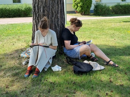 Två ungdomar sitter på en gräsmatta  lutade mot ett träd och skriver i anteckningsblock samt tittar på en surfplatta