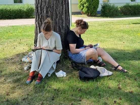 Två ungdomar sitter på en gräsmatta lutade mot ett träd och skriver i anteckningsblock samt tittar på en surfplatta