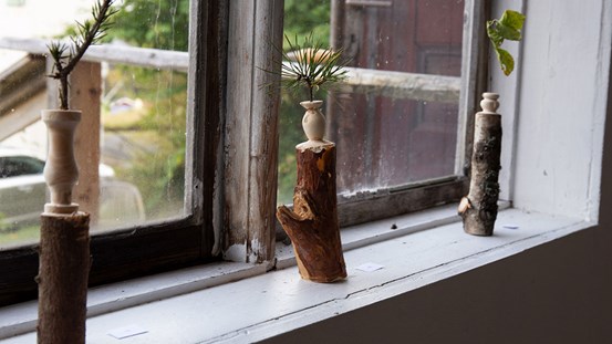 Foto. Tre träskulpturer, en del av en trädstam/gren där den övre delen har svarvats i form av en vas. I varje vas är en kvist nedstoppad.  De står på rad på fönsterbrädan i ett fönster. Ur fokus utanför fönstret syns en del av en dörr och grön natur. 