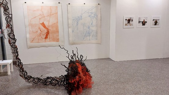 Foto av galleri Lokomotivs monter. Ett rum med en vit vägg , eller skärm. Närmast till vänster, på det grå golvet, ligger en textil skulptur. Den är virkad av järntråd och röd och svart sisal. Verket hänger från taket. Den långa smala delen kommer ner i fotots övre vänstra hörn och går ner till golvet där den sedan fortsätter åt höger en bit. Där blir den större och breder ut sig över golvet som ett domedagsmoln. Bakom på väggen syns två stora grafiktryck, ett i röda toner och ett i blåa. Längre bort, till höger på väggen syns tre stycken foton, det ser ut som det är människor i ett landskap.