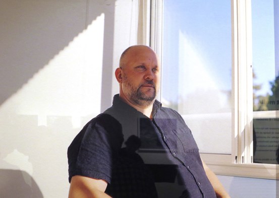 Foto, ett porträtt i halvfigur av en sittande man. Han har kort skägg, kalt huvud och en mörkblå kortärmad skjorta. Han sitter framför en vit vägg till vänster i bild och solen faller in genom ett fönster till höger.