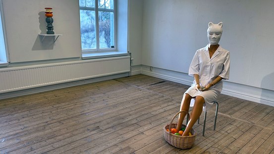 Ett rum med trägolv och vita väggar, ett fönster på den vänstra väggen. Till höger i bild, på en pall, sitter en skyltdocka iförd en vit mask, vit sjukhusrock och med fötterna i en korg med frukt. Händerna håller en liten hög med aska.