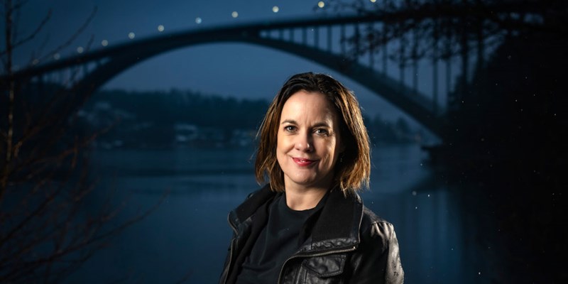 Porträttfoto av Elin Holmberg. Kvinna med mörkbrunt hår, skinnjacka. Sandöbron i bakgrunden mot en mörkblå kvällshimmel.
