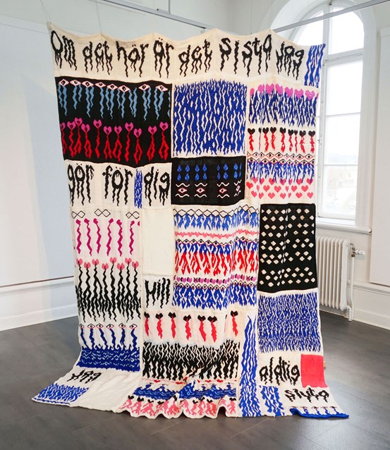 Ett stort täcke hänger från taket, i bakgrunden vita väggar och ett fönster. Beskrivning av verket finns i brödtext. 