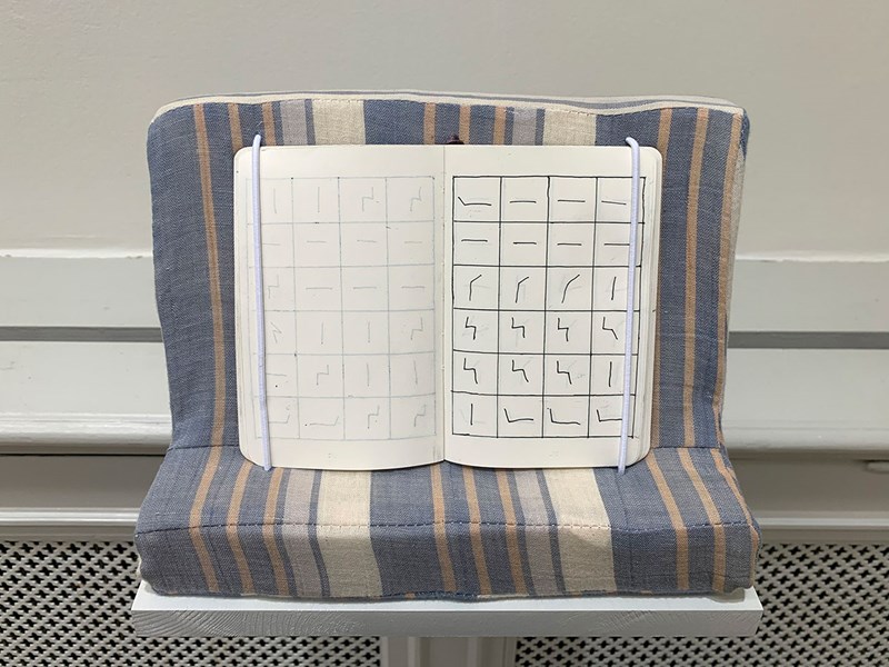 Fastsatt på väggen är en hylla liknande en soffa, ett L-format ställ klädd i ett randigt tyg. På ”soffan” står en bok uppslagen. Det är vita blad som har 4 x 6 rutor, i varje ruta är ett tecken. Allt ser ut att vara tecknat i svart.