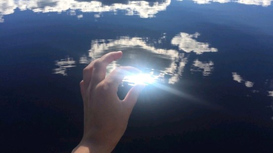 Foto av en hand framför en spegling av himlen och solen i vatten. Tummen och pekfingret hålls runt solen. 