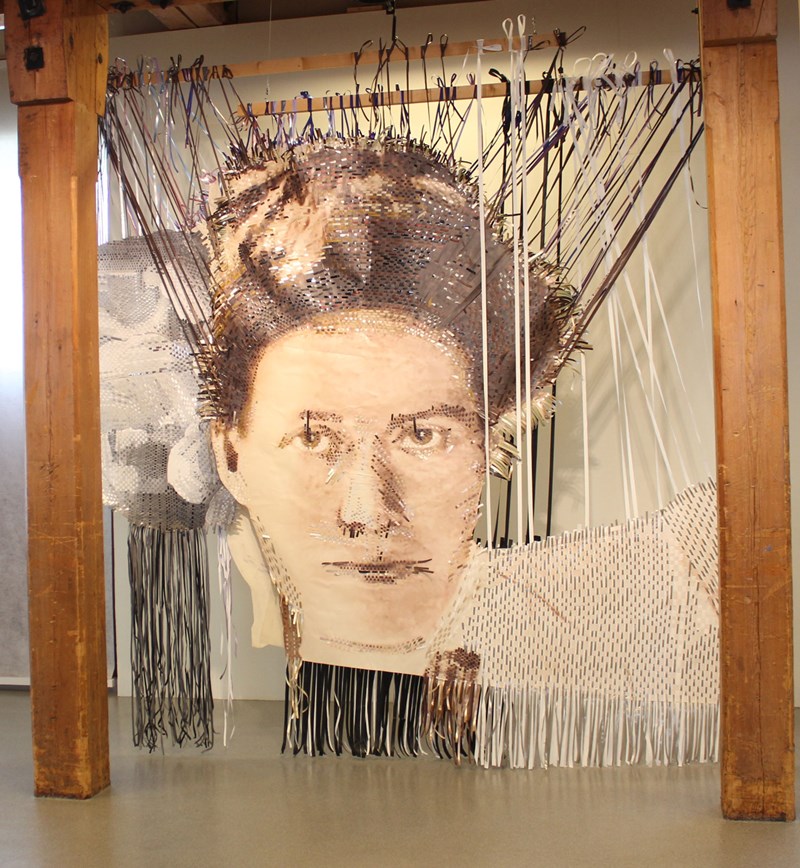 Mellan två träpelare sitter Kristina Müntzings verk upphängt. Det är ett stort utskrivet porträttfoto i sepia av en kvinna. I utskriften är plastband inflätade. En del av banden som är invävda i bilden är uppspända i träribbor ovanför den. 