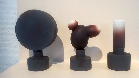 Tre skulpturala objekt står på rad på en ljus yta. Det är olika geometriska former på varsin cylindrisk fot. De är alla svarta eller mörka i färgen, förutom längst upp där det mörka tonar över till ljust och vitt. 