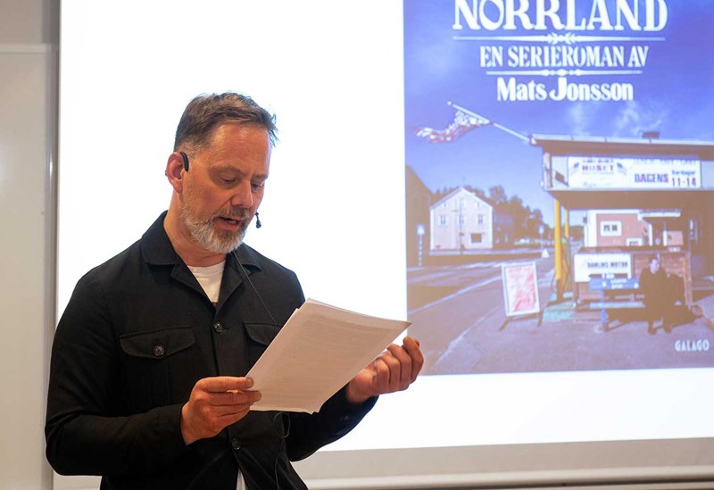 Mats Jonsson står framför projektorduk som visar en omslaget på hans bok. Han är iklädd en svart skjorta och tittar ner på papperet han håller i handen. 