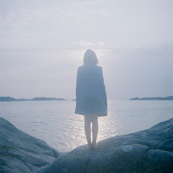 Fotografi i blå, disiga toner. En person står på klippor med ryggen mot betraktaren och vänd ut mot havet. Där möter havet himlen med avlägsna klippor. 