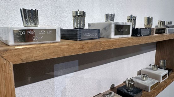 En trähylla med två hyllplan på en vit vägg. På den står rader med socklar med ljus i metall. 