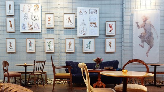 På ett grått galler som täcker hela den stora grå väggen hänger målningar och teckningar av människor, kroppar, gestalter. Framför väggen med bilder står grupper med stolar och bord i varierande modeller och även en mörk blå soffa.
