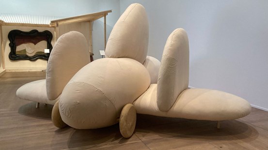 En skulptur, ett flygplan i runda bulliga former klädd i beige textilmaterial och med trähjul. 