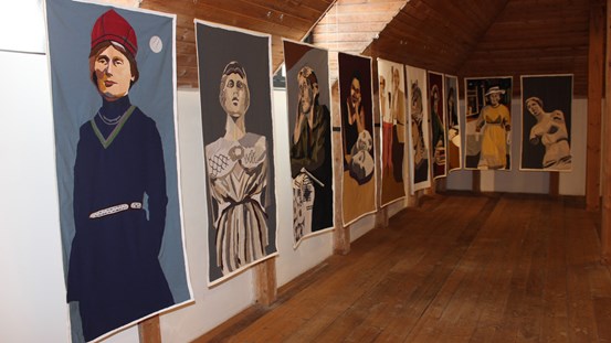 I ett rum med trägolv och snedtak hänger åtta stora bilder. Betraktaren ser bilderna till vänster på fotot, de sträcker sig bortåt till väggens hörn för att sedan fortsätta på nästa vägg. Bilderna är textila kollage och föreställer historiska kvinnor. 