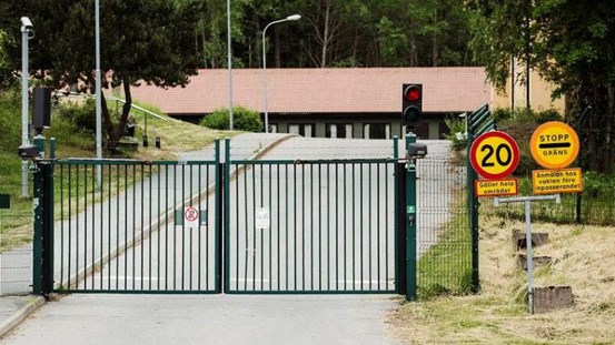Fotografi som visar en stängd grind med skylten STOPP-GRÄNS på högersidan. Infarten till FRA. Vägen leder uppför en kulle, i bakgrunden syns en byggnad.
