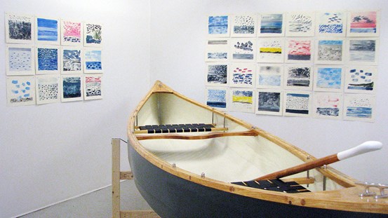 Utställningsrum som visar två väggar fylld med kvadratiska monotypier i blått, rosa och gult. På golvet har en kanot placerats.