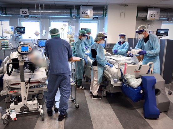 Personal i arbete på intensivvårdsavdelning. Fem personer i skyddsutrustning står samlade runt en patient i en säng.