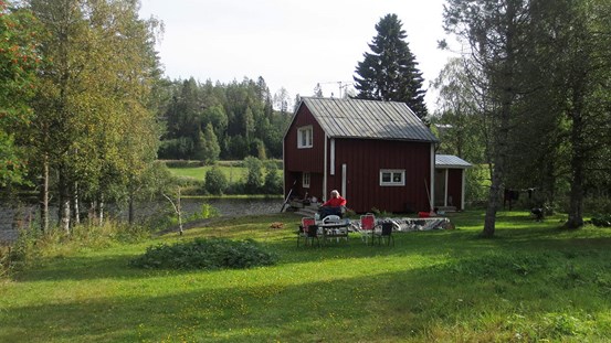 Fotografi av stipendiestugan i Grundsunda en sommardag i augusti. En röd stuga med vita knutar omgiven av några träd. I bakgrunden ett vattendrag och skog.