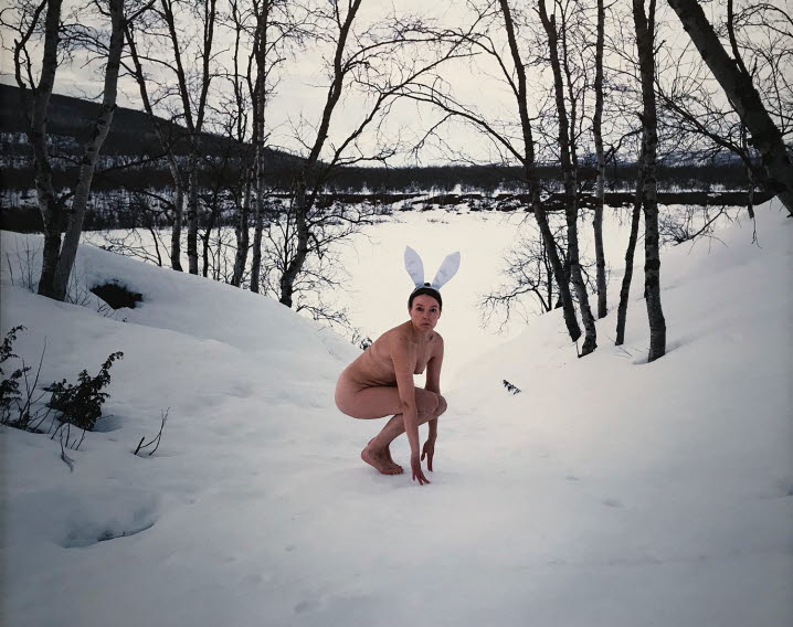 Foto av en naken kvinna, med vita kaninöron, sittandes på huk likt en kanin eller hare i en snöigt landskap. Marja Helander, Blickande mot kvällen. ©Marja Helander.