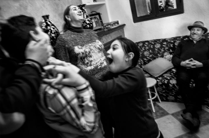 Åke Ericsons svartvita fotografi av romer i Calanas, Spanien. En hemmiljö med glada barn som busar. En äldre man sitter nöjt leende i soffan och en kvinna skrattar.