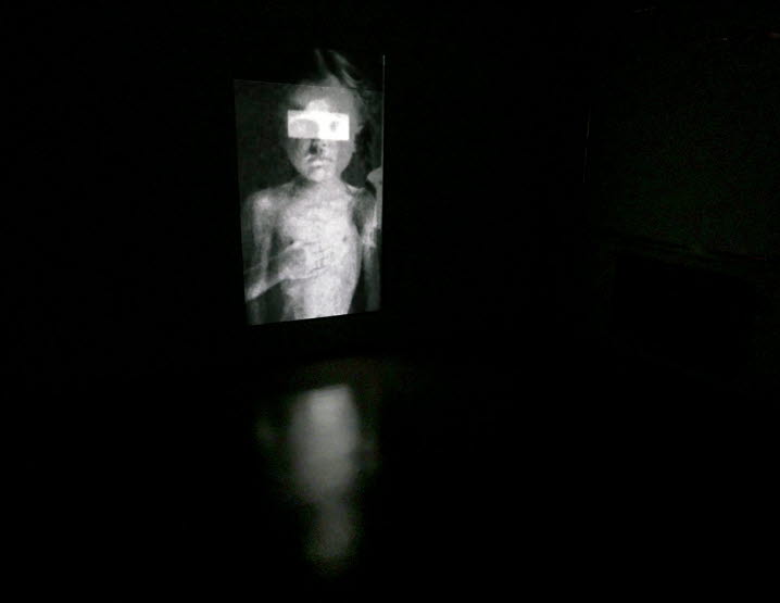 Dokumentation av Magnus Wallins bildspelet ”Unnamed” som projiceras mot en vägg i stående format. Stillbilden visar ett barn, med högerhanden mot bröstet och en ljus rektangel över ögonen.