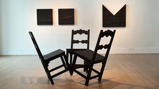 Utställningsvy från konsthallen i Luleå. På golvet står en skulptur bestående av tre svarta stolar som vinklas bakåt, sammanfogade i de två främre stolsbenen. På väggen hänger tre trä verk av mörkt trä. 